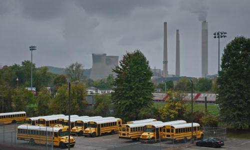 A line of school buses in front of smoke stacks.